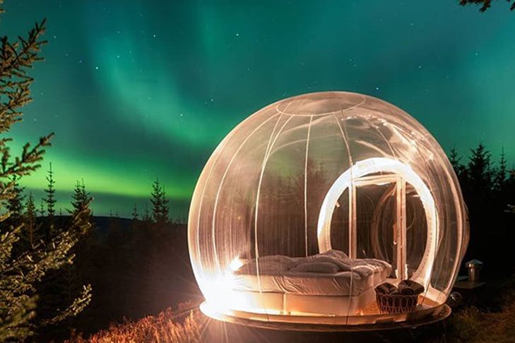 A clear bubble with a bed inside underneath the Northern Lights.